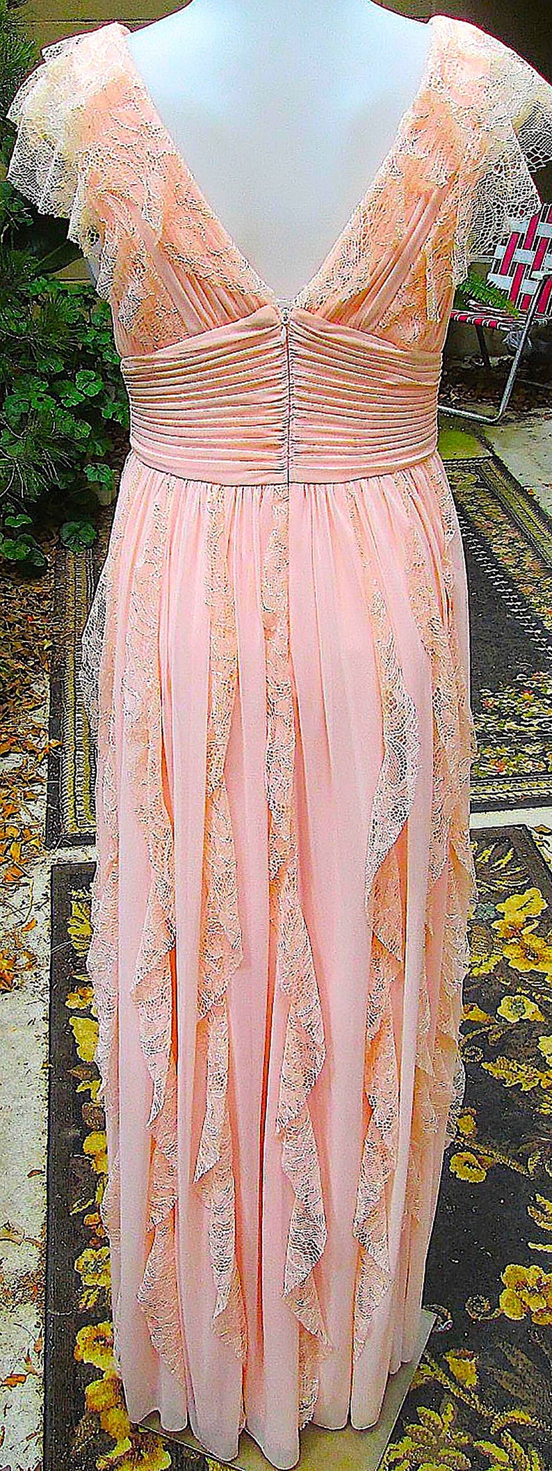 K&ouml;nnte beinhalten: Ein bodenlanges, rosa Vintage-Kleid mit zarten Spitzenbes&auml;tzen. Das Kleid hat einen V-Ausschnitt, eine betonte Taille und flie&szlig;ende Rockbahnen mit Spitzenakzenten. Das Kleid ist aus einem weichen, flie&szlig;enden Stoff gefertigt.