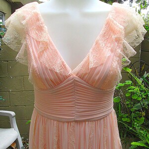 K&ouml;nnte beinhalten: Ein hellrosa Abendkleid mit V-Ausschnitt und zarten Spitzendetails. Das Kleid hat ein figurbetontes Oberteil mit plissierten Akzenten und einen flie&szlig;enden Rock. Die Schultern haben ger&uuml;schte Spitzen&auml;rmel.
