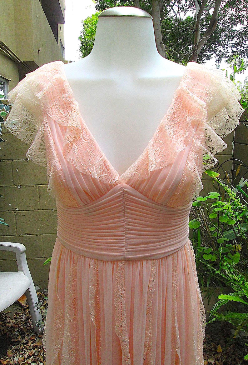 K&ouml;nnte beinhalten: Ein hellrosa Abendkleid mit V-Ausschnitt und zarten Spitzendetails. Das Kleid hat ein figurbetontes Oberteil mit plissierten Akzenten und einen flie&szlig;enden Rock. Die Schultern haben ger&uuml;schte Spitzen&auml;rmel.
