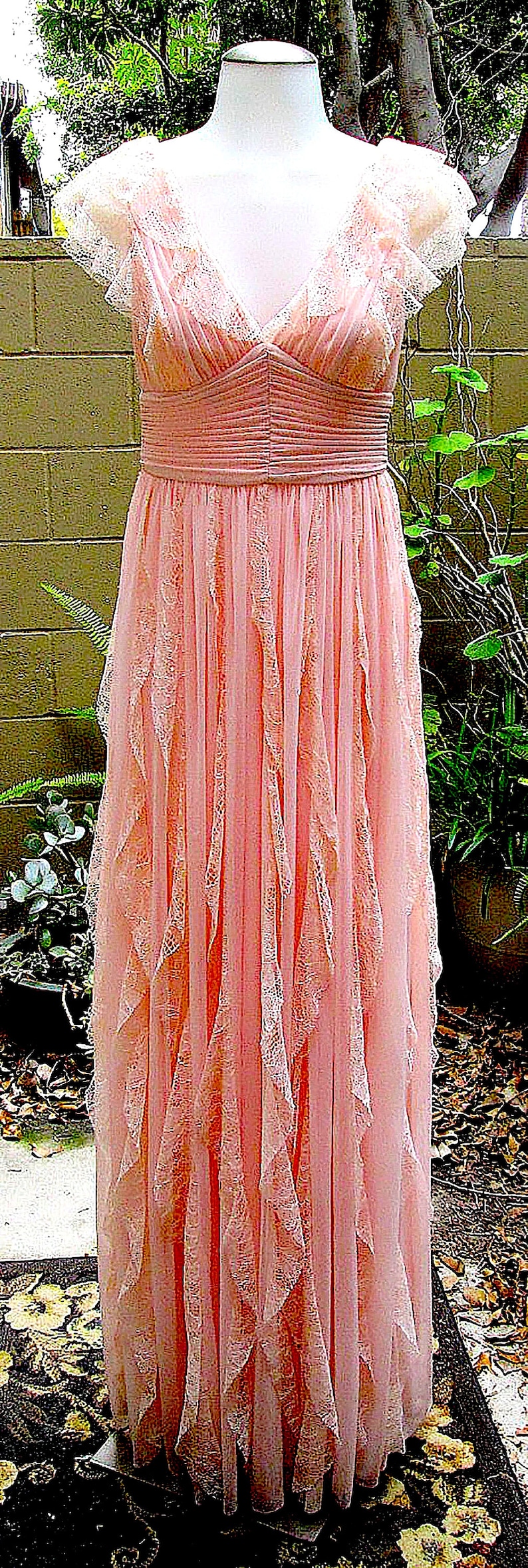 May include: A pink dress with a lace overlay and a v-neckline. The dress is pleated and has a ruffled skirt. The dress is displayed on a mannequin against a brown brick wall background.