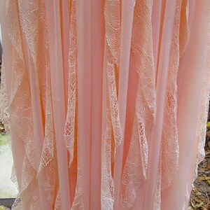 K&ouml;nnte beinhalten: Ein flie&szlig;endes, hellrosa Kleid mit zarten Spitzenbesatz. Das Kleid hat vertikale Bahnen aus durchsichtigem Stoff, wobei Spitzenelemente &uuml;ber die Vorderseite fallen. Der Gesamteffekt ist weich und romantisch.