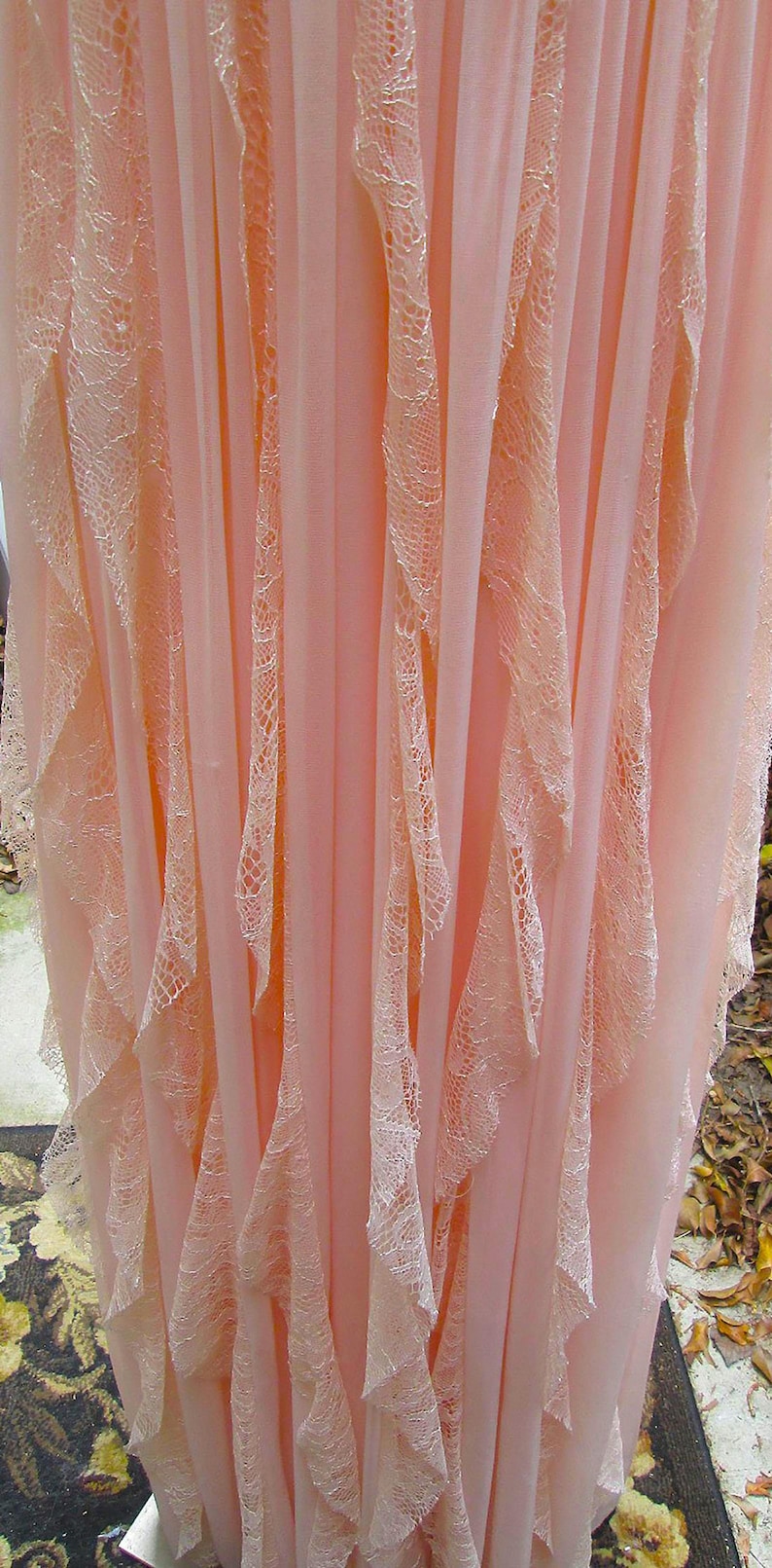 K&ouml;nnte beinhalten: Ein flie&szlig;endes, hellrosa Kleid mit zarten Spitzenbesatz. Das Kleid hat vertikale Bahnen aus durchsichtigem Stoff, wobei Spitzenelemente &uuml;ber die Vorderseite fallen. Der Gesamteffekt ist weich und romantisch.