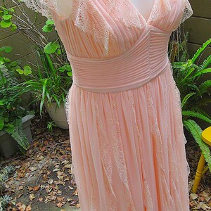 K&ouml;nnte beinhalten: Ein hellrosa Abendkleid mit V-Ausschnitt und kurzen, ger&uuml;schten &Auml;rmeln. Das Kleid hat zarte Spitzentrimm und ein plissiertes Oberteil mit flie&szlig;endem Rock. Das Kleid wird auf einer Schaufensterpuppe pr&auml;sentiert.