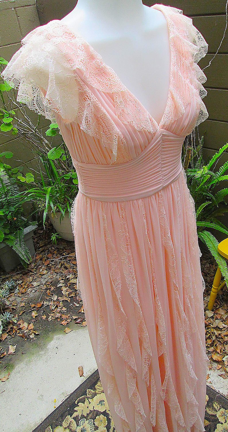 K&ouml;nnte beinhalten: Ein hellrosa Abendkleid mit V-Ausschnitt und kurzen, ger&uuml;schten &Auml;rmeln. Das Kleid hat zarte Spitzentrimm und ein plissiertes Oberteil mit flie&szlig;endem Rock. Das Kleid wird auf einer Schaufensterpuppe pr&auml;sentiert.