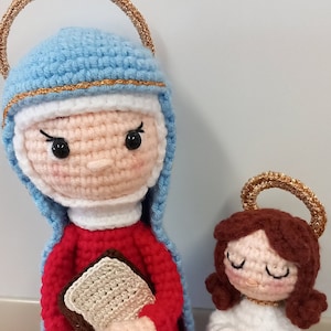 May include: Crocheted figures of the Virgin Mary and a small angel. The Virgin Mary is wearing a blue robe with a gold halo and holding a book. The angel is wearing a white robe with a gold halo and has brown hair.