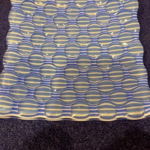 May include: Square glass dish with a wavy edge. The dish features a blue and white striped pattern with raised, rounded sections. The dish is on a dark blue surface.