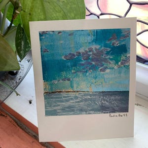 May include: A painting depicting a seascape with a blue sky and white clouds. The water is a light blue and the horizon is a thin line of yellow. The painting is signed "Pauline Fox 123" in the bottom right corner.