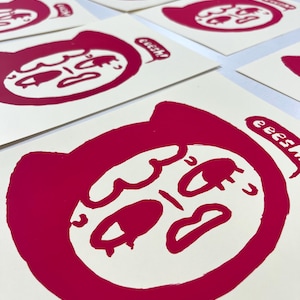 Puede incluir: Múltiples impresiones de una cara roja estilizada con una expresión angustiada y la palabra "eeesh" en una burbuja de diálogo. Las impresiones están sobre papel blanquecino, creando un contraste audaz. La obra de arte tiene un estilo gráfico moderno.