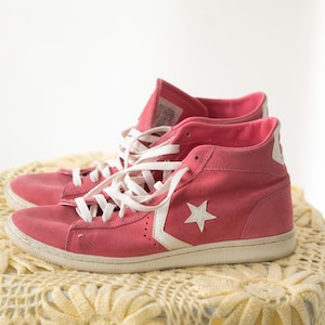 May include: A pair of pink high-top sneakers with white laces and a white star logo on the side. The shoes are placed on a yellow crocheted table cover. The sneakers have a white sole and are made of a suede-like material.