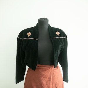 May include: Black velvet jacket with gold trim and decorative floral accents. The jacket is displayed on a mannequin with a rust-colored skirt. The jacket has long sleeves and a cropped design, suitable for vintage or retro fashion.