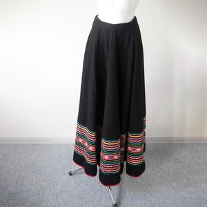May include: A floor-length black skirt featuring a wide, flared silhouette and a high waistband. The hem is decorated with multiple horizontal bands of colorful embroidery in shades of red, orange, green, blue, and white.