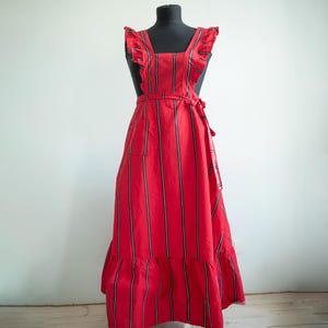May include: A red apron with vertical black and white stripes, displayed on a black mannequin. The apron features ruffled shoulder straps, a waist tie, a front pocket, and a ruffled hemline. The apron is a full-length design.