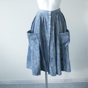 Può includere: Una gonna blu al ginocchio con una texture screziata effetto lavaggio acido. La gonna presenta una vita alta, una fila verticale di bottoni bianchi sul davanti e due grandi tasche applicate sui lati. Il tessuto ha un design plissettato.