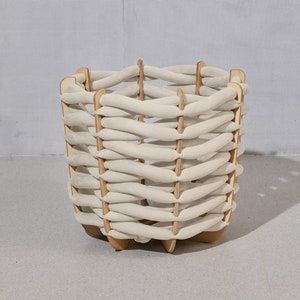 May include: A square, woven basket with a light beige fabric and wooden frame. The basket has a unique design with horizontal fabric strips woven between vertical wooden supports. The basket is sitting on a gray surface.