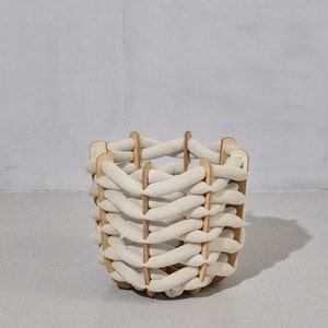 May include: A woven basket with a unique design. The basket features a cream-colored, braided exterior supported by wooden vertical slats. The basket is cylindrical and sits on a light gray surface, with a light gray background.