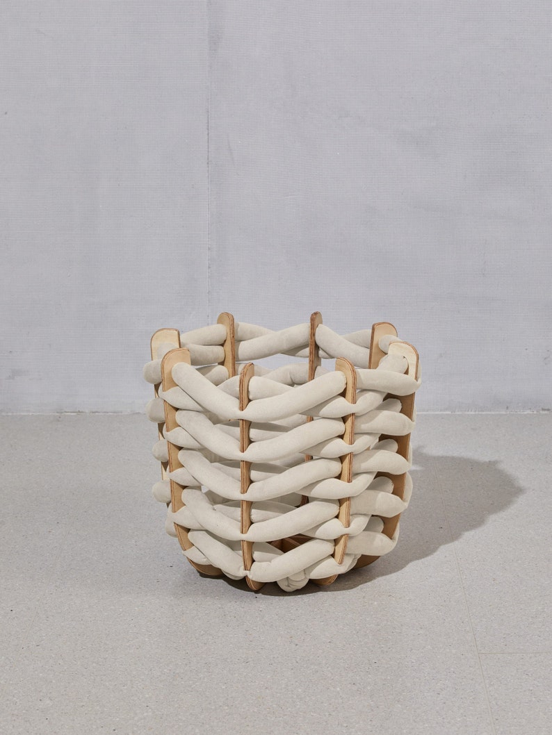 May include: A woven basket with a unique design. The basket features a cream-colored, braided exterior supported by wooden vertical slats. The basket is cylindrical and sits on a light gray surface, with a light gray background.