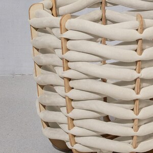 May include: A woven basket with a unique design. The basket features a light beige, braided pattern made of soft material, supported by a wooden frame. The wooden frame is a light brown color. The basket has a rounded shape.