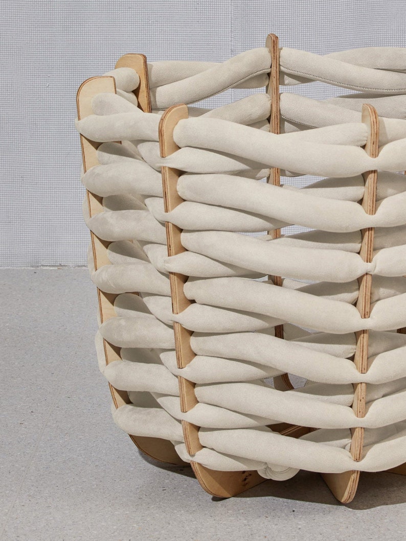 May include: A woven basket with a unique design. The basket features a light beige, braided pattern made of soft material, supported by a wooden frame. The wooden frame is a light brown color. The basket has a rounded shape.