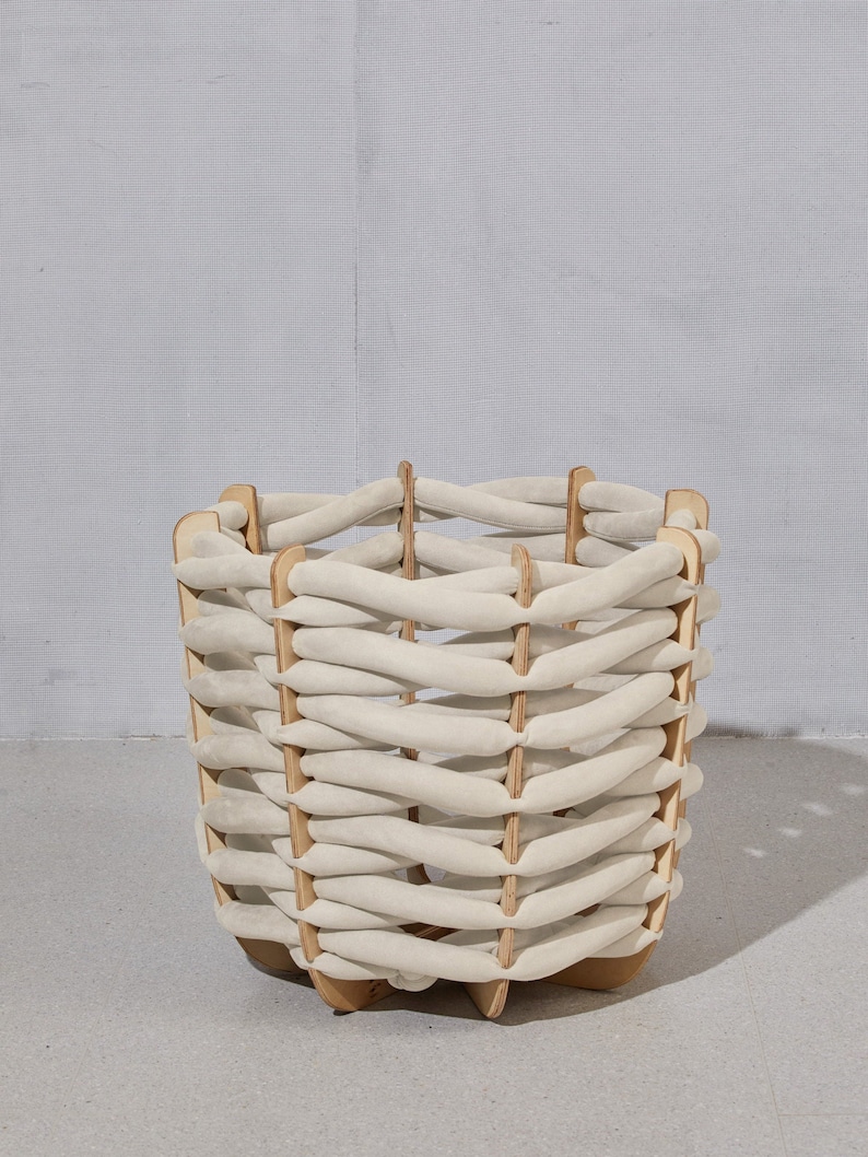 May include: A square, woven basket with a light beige fabric and wooden frame. The basket has a unique design with horizontal fabric strips woven between vertical wooden supports. The basket is sitting on a gray surface.