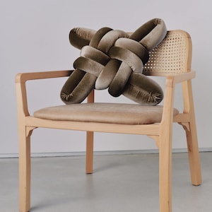 Pode incluir: Uma almofada decorativa em forma de nó, em tons de verde oliva e bege, repousa sobre uma cadeira de madeira clara com encosto tecido e assento bege. O design da almofada apresenta uma forma de nó, adicionando um toque único à estética da cadeira.