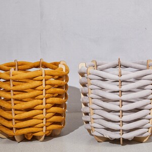 Pode incluir: Duas cestas decorativas com design entran&ccedil;ado. Uma &eacute; amarelo mostarda, a outra cinza claro. Ambas t&ecirc;m uma estrutura de madeira. As cestas est&atilde;o sobre um fundo neutro. Adequadas para armazenamento.