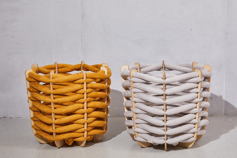 Pode incluir: Duas cestas decorativas com design entran&ccedil;ado. Uma &eacute; amarelo mostarda, a outra cinza claro. Ambas t&ecirc;m uma estrutura de madeira. As cestas est&atilde;o sobre um fundo neutro. Adequadas para armazenamento.