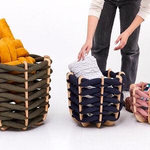 May include: Three decorative woven storage baskets in olive green, navy blue, and pink. Each basket has a wooden frame and is filled with items. The olive green basket contains a mustard-colored cushion. The navy basket holds a gray blanket. The pink basket has books.