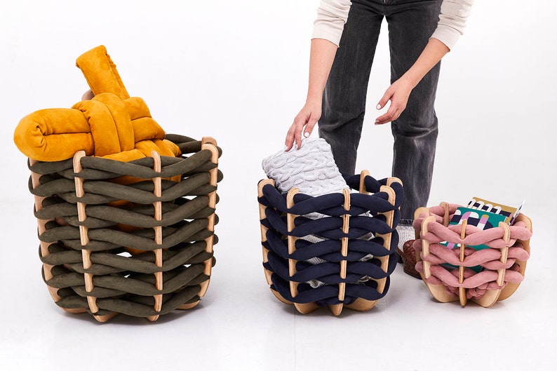 May include: Three decorative woven storage baskets in olive green, navy blue, and pink. Each basket has a wooden frame and is filled with items. The olive green basket contains a mustard-colored cushion. The navy basket holds a gray blanket. The pink basket has books.