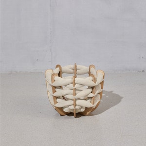 May include: A decorative basket with a unique design. The basket features a wooden frame with interwoven cream-colored fabric strips. The basket is a light brown color and is sitting on a gray surface.