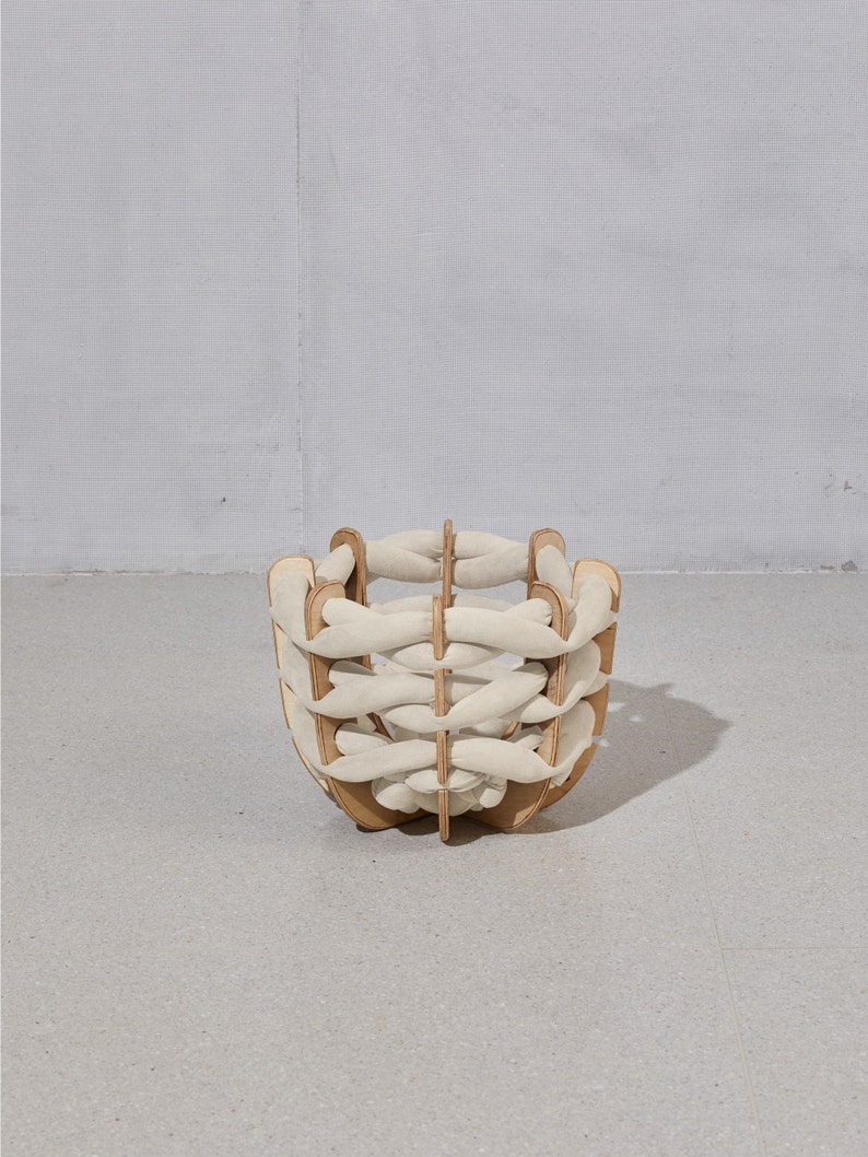 May include: A decorative basket with a unique design. The basket features a wooden frame with interwoven cream-colored fabric strips. The basket is a light brown color and is sitting on a gray surface.