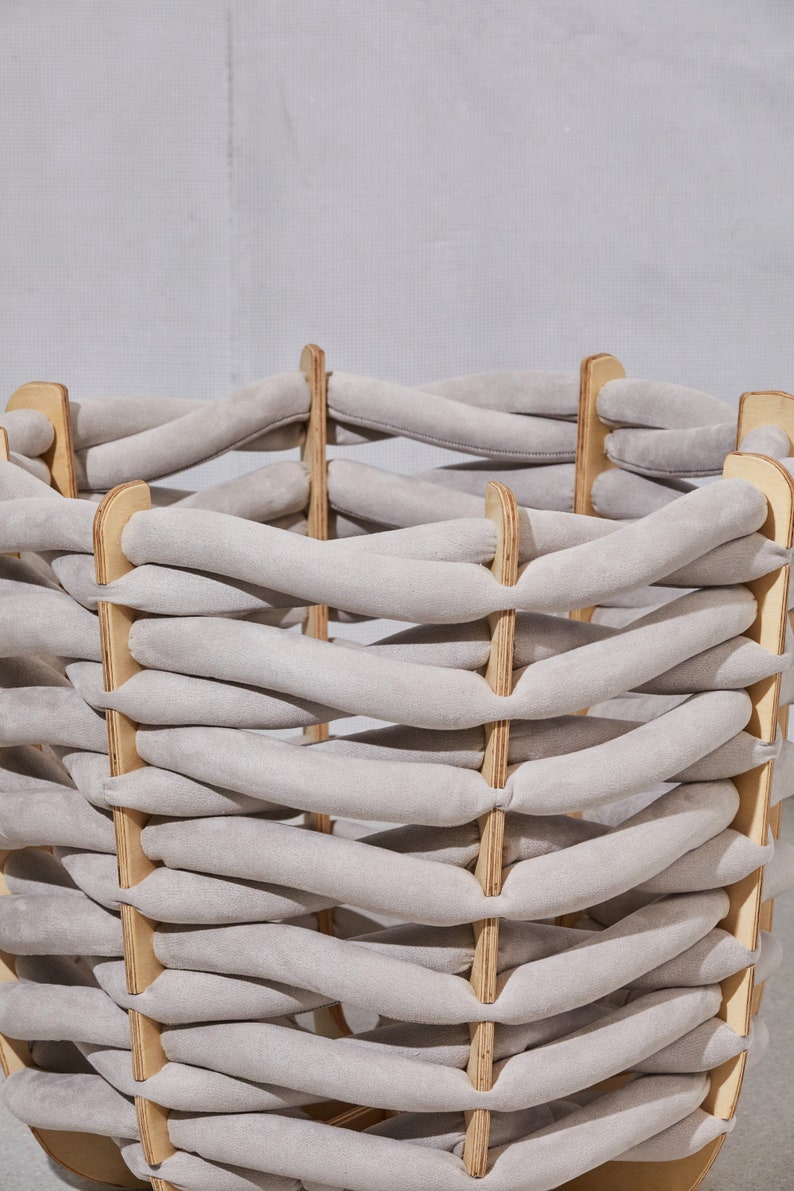 Pode incluir: Um cesto tecido com um design de tecido cinza claro. O cesto &eacute; constru&iacute;do com suportes de madeira e tiras de tecido acolchoadas entrela&ccedil;adas. A forma geral &eacute; cil&iacute;ndrica, com uma base resistente e uma parte superior aberta. A paleta de cores &eacute; neutra.