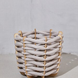 Pode incluir: Uma cesta tecida com um design de tecido cinza claro. A cesta tem uma estrutura de madeira e uma forma arredondada. O padr&atilde;o tecido &eacute; feito de tecido grosso e macio, cinza claro. A cesta est&aacute; em uma superf&iacute;cie cinza claro.