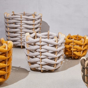 May include: A collection of woven baskets in various sizes and colors. The baskets feature a wooden frame with a woven fabric design in shades of gray, mustard yellow, and olive green. The baskets are arranged on a light gray surface.