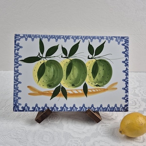May include: Rectangular ceramic serving dish with a hand-painted design of three green apples with yellow highlights and dark green leaves. The dish has a blue and white border and is displayed on a wooden stand. A yellow lemon sits to the side.