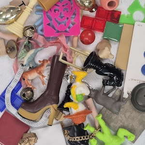 May include: A collection of small, colorful objects including a brown platform boot, a red tile, a green plastic figure, and a card of blue buttons. Other items include a small cow figurine, bells, and wooden letter tiles.