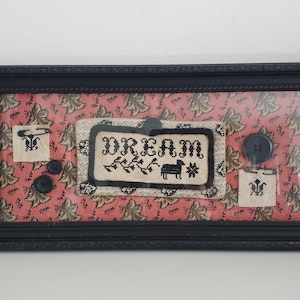 May include: A framed sampler with a pink and brown floral pattern. The sampler features a cross-stitched design that says "DREAM" with a small black sheep and a star. There are also three small cross-stitched squares with black designs and two black buttons.