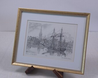 May include: A framed monochrome print of a sailing ship in a harbour, with buildings and a church spire in the background. The frame is gold-coloured, and the print is displayed on a wooden easel.