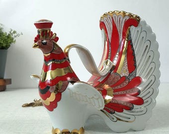May include: A porcelain bird-shaped decanter featuring a white base with vibrant red, black, and gold decorative patterns. The vessel includes a removable stopper on the head, a curved handle, and an elaborate, fanned tail design with gold trim.
