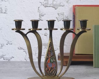 May include: A brass candelabra featuring five candle holders with dark metal cups. The central stem displays a colourful enamel inlay of a stylised deer. The base is oval-shaped, and the arms curve outward in a symmetrical, fluid design.