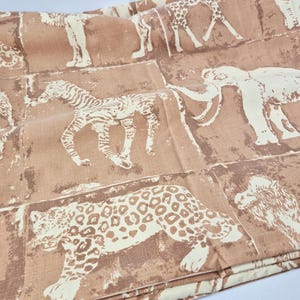 May include: Tan fabric featuring a repeating pattern of safari animals in off-white. The design includes zebras, giraffes, elephants, and leopards. The fabric is folded, showing multiple panels of the animal print.