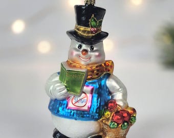 May include: A glass snowman ornament wearing a black top hat and a blue vest, holding a green book labelled DIABETES and a basket of red fruit. The snowman has a white body, a gold patterned scarf, and a sign on the vest that says NO SUGAR.