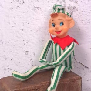 May include: A small elf doll with a peach-colored face, blue eyes, and a smiling expression. The doll wears a green and white vertically striped fabric suit with a matching pointed hat and a bright red collar piece around the neck area.