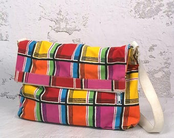 May include: A rectangular fabric messenger bag featuring a vibrant multi-coloured plaid pattern in red, yellow, orange, pink, green, and blue squares separated by black lines. The bag includes a fold-over flap, a white strap, and a side zipper.