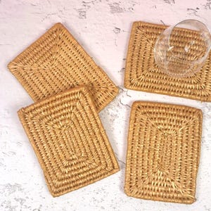 May include: A set of three rectangular woven rattan coasters with a light tan natural finish. Each coaster features a tight, concentric braided pattern. One clear glass tumbler sits on the top right coaster, showcasing the woven texture.