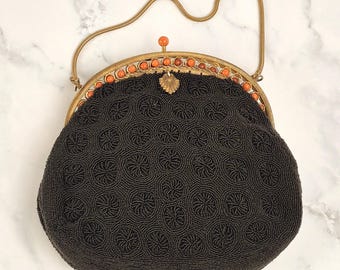 May include: A black beaded handbag featuring a repeating circular pattern. The bag has a gold-toned metal frame adorned with small orange beads along the top edge, a shell-shaped clasp, and a thin gold-toned metal chain handle for carrying.