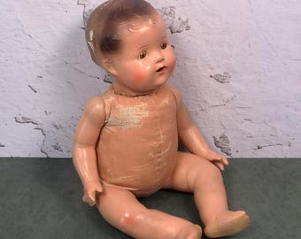 May include: A vintage doll with a moulded composition head featuring painted brown hair, brown eyes, and rosy cheeks. The doll has a soft, fabric-covered torso with composition arms and legs, showing visible signs of wear and ageing on the surface.