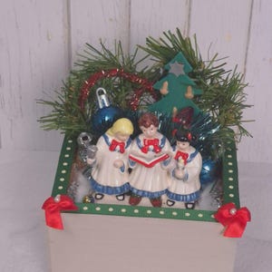 May include: A decorative box featuring three ceramic choir figures in white robes with blue trim and red bows, holding songbooks. The arrangement includes green pine branches, a small green tree cutout, blue ornaments, and red ribbon bows.