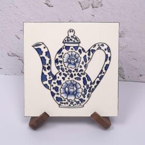 May include: A square ceramic tile featuring a central illustration of a teapot with a blue and white mottled pattern and two large floral designs. The teapot has a lid, a curved spout, and a handle, all outlined in black on a cream background.