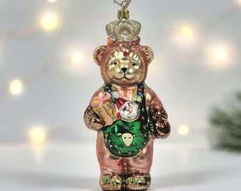 May include: A glass Christmas ornament shaped like a bear wearing a green apron with a reindeer graphic and a Santa face. The bear is metallic copper in colour, holds a pink gift, and has a small crown on its head with a star-shaped metal hanger.