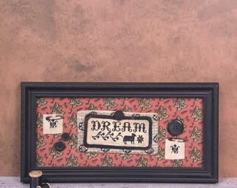 May include: A rectangular black frame containing a red fabric background with a repeating leaf pattern. Centered is a cream-coloured fabric patch with the black embroidered text "DREAM" and a sheep icon, accented by black buttons and pins.